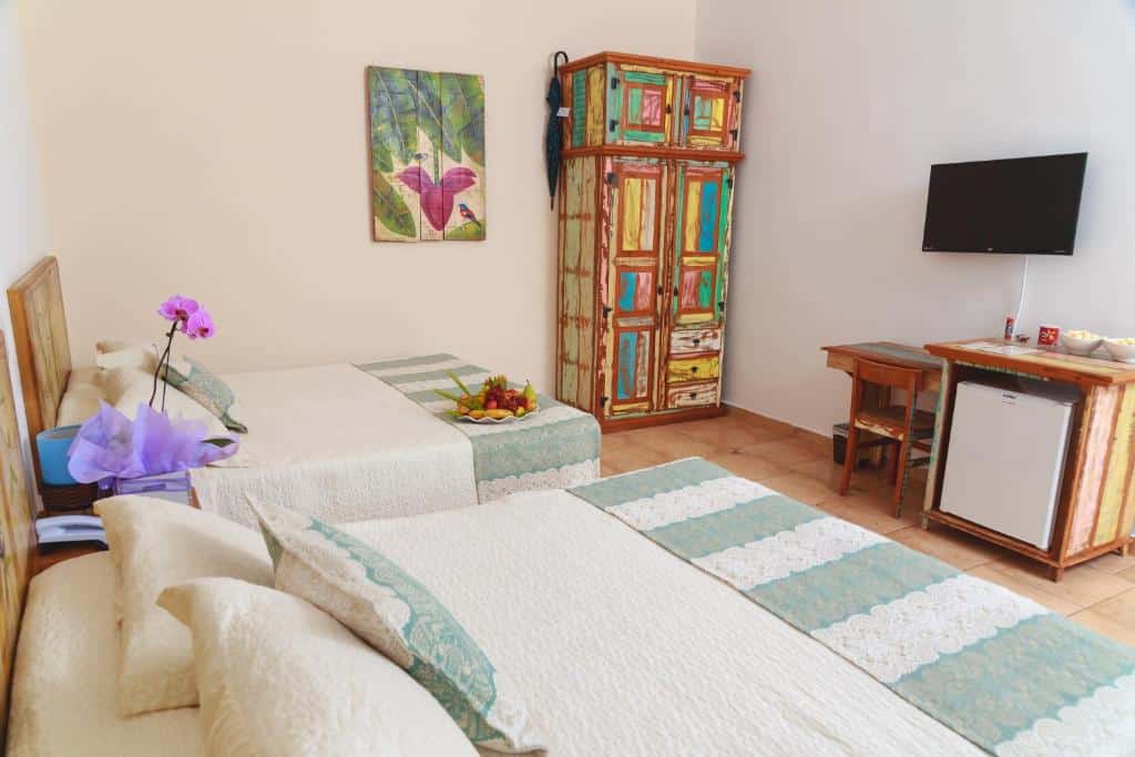 Quarto do Hotel Fazenda Hípica Atibaia com duas camas de casal do lado esquerdo do quarto em frente as camas mesa de trabalho de madeira com cadeira , TV presa na parede e um frigobar do lado da mesa.