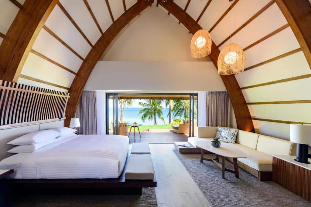 Quarto do Fiji Marriott Resort Momi Bay com cama de casal ampla, em frente a cama sofá cor bege com mesinha de centro redonda a frente, do lado esquerdo portas de vidro com varanda e vista para o mar.