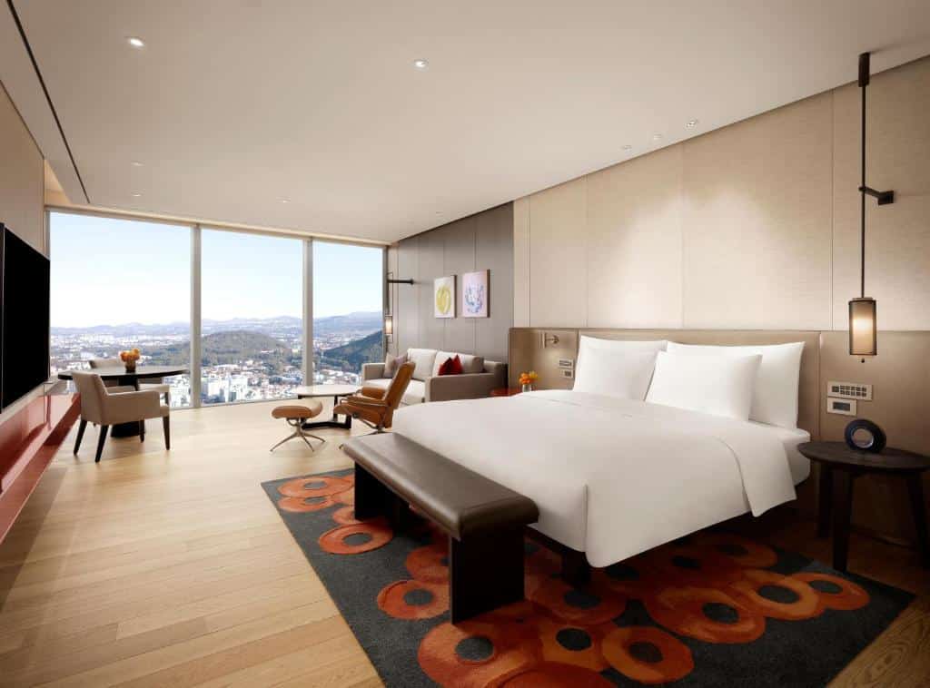 Quarto amplo do Grand Hyatt Jeju com janela panorâmica com vista para a cidade e montanhas, televisão, sofá com almofadas, poltronas, cama de casal e uma tapete preto com círculos redondos, para representar hotéis na Coreia do Sul