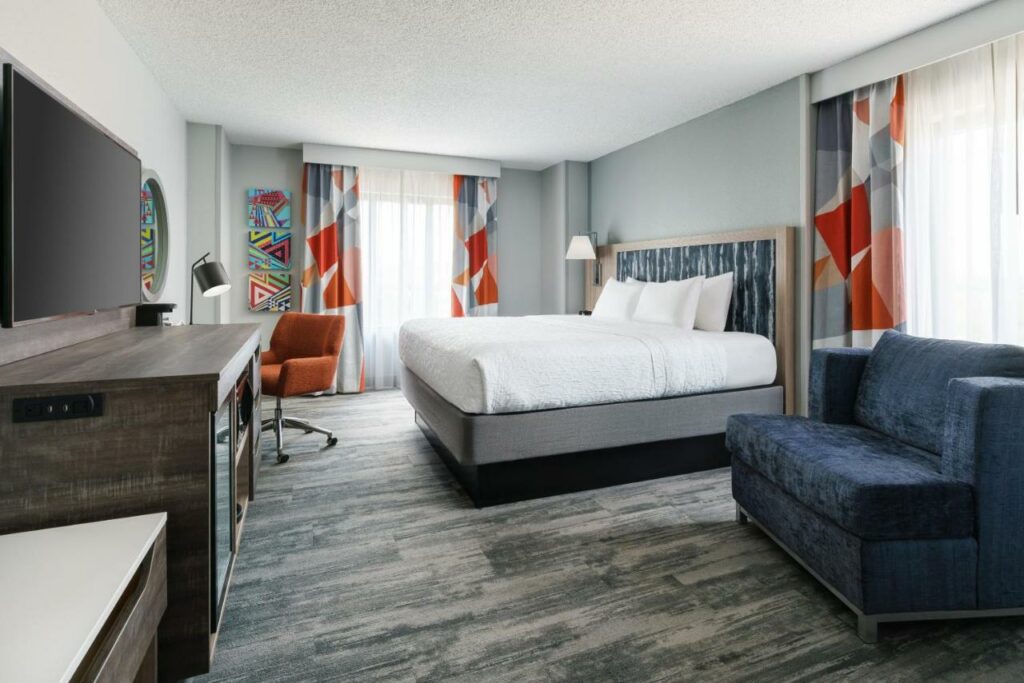 Quarto do Hampton Inn & Suites Tampa Ybor City Downtown com uma cama de casal, duas janelas com cortinas, chão de carpete, uma poltrona, uma mesa de escritório com uma cadeira, para representar hotéis perto do Busch Gardens