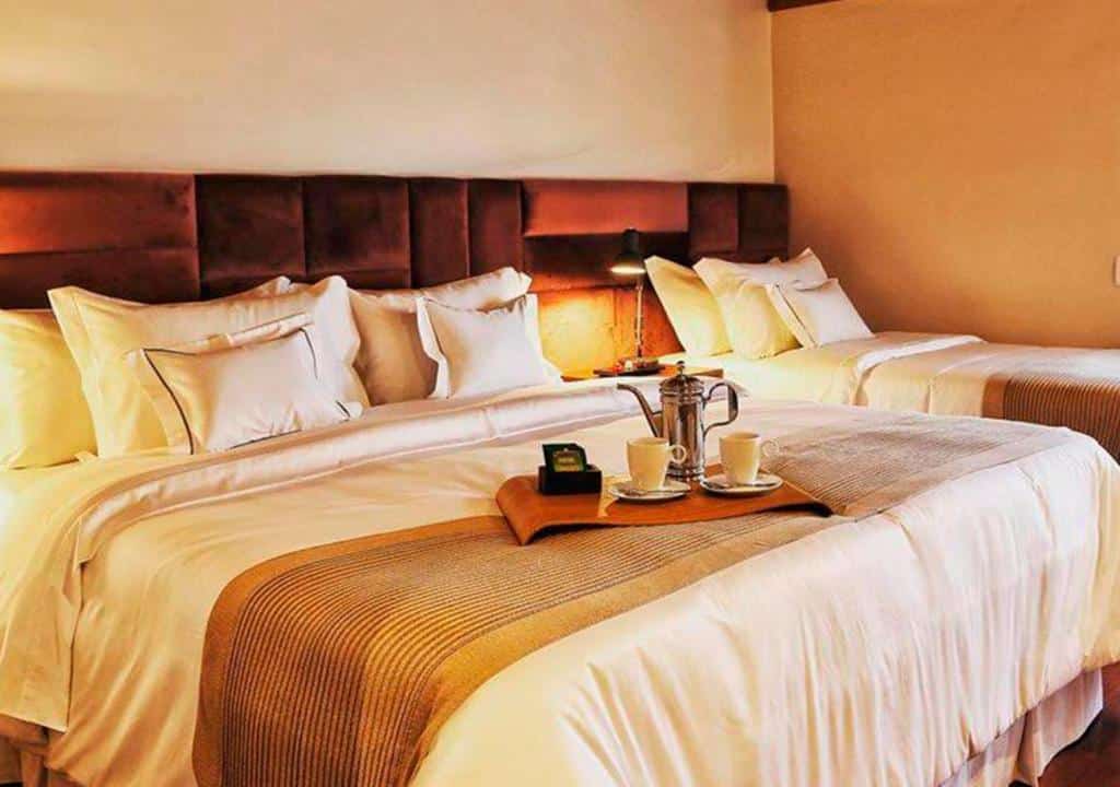 Quarto do Hotel Boutique QUEBRA-NOZ com uma cama de casal com uma bandeja com duas xícaras e um bule, e do lado direito há mais uma cama de solteiro e entre as camas há uma luminária