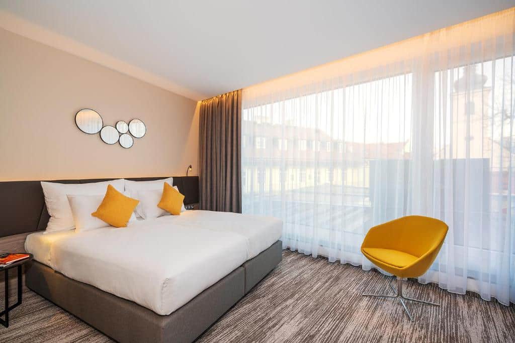 Quarto do Hotel CUBE Prague com uma janela ampla de vidro com cortinas, uma cama de casal com travesseiros e almofadas, uma poltrona e um chão com carpete marro e branco