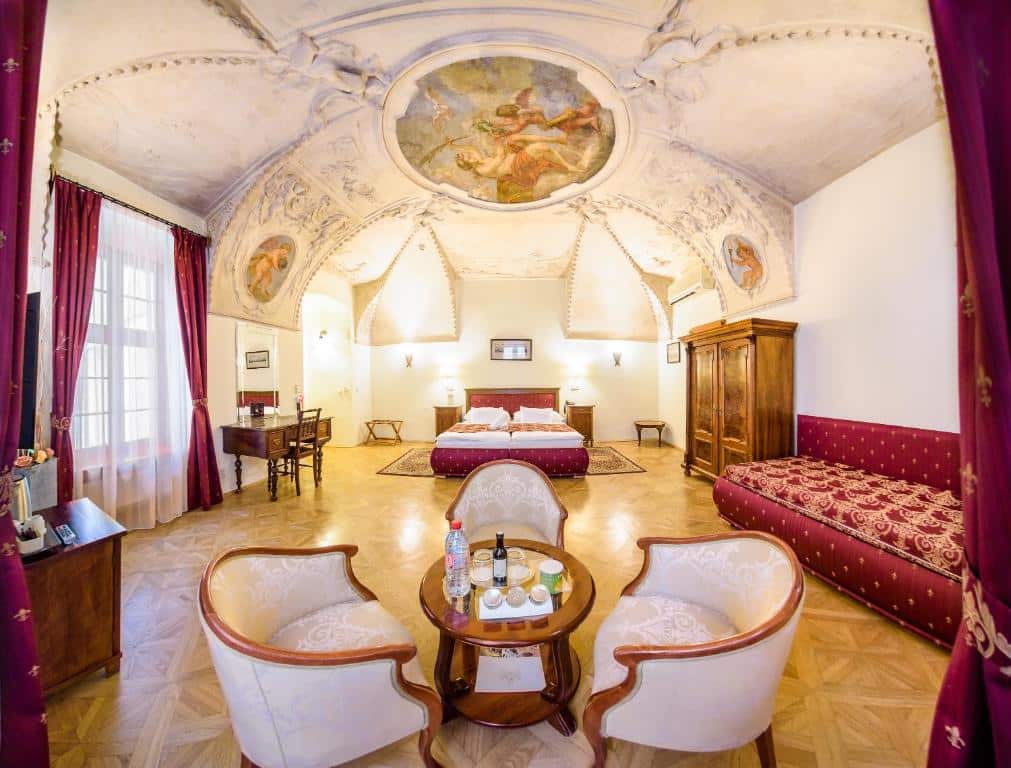 Quarto do Hotel Elite Prague com espaço amplo, o teto é renascentista com desenhos de anjos, há um sofá vermelho, uma cama de casal, um armário com duas portas e uma mesa redonda com três poltronas ao redor