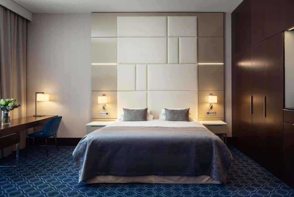 Quarto do Hotel KINGS COURT com uma carpete azul marinho, cama de casal, duas mesinhas de cabeceira com luminárias, uma mesa de trabalho com uma cadeira e uma janela com cortinas