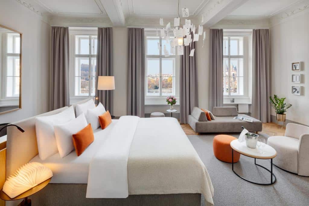 Quarto espaçoso do Hotel Leonardo & Bookquet Prague com três janelas com cortinas, um lustre, uma cama de casal, um pequeno sofá e uma poltrona, com um tapete cinza