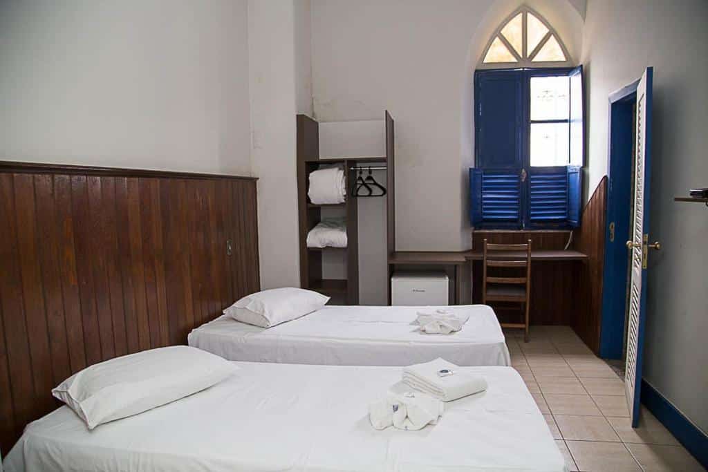 quarto antigo do Hotel Providencia com duas camas de solteiro, um armário sem porta, um frigobar e uma pequena mesa de madeira com cadeira. A janela e a porta estão pintadas em azul royal