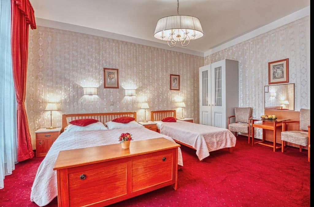Quarto amplo do Hotel Salvator com uma janela com cortinas, uma cama de casal, uma cama de solteiro, um pequeno armário branco com duas portas, duas poltronas, um pequena penteadeira com espelho, tudo decorado em branco e vermelho