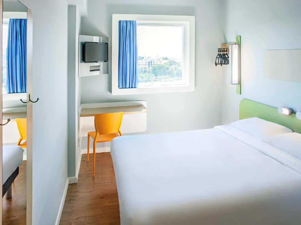 Quarto do hotel com cama branca, paredes brancas, janela de vidro, uma escrivaninha, cadeira amarela e uma tv, ilustrando post hotéis Ibis no Rio de Janeiro.