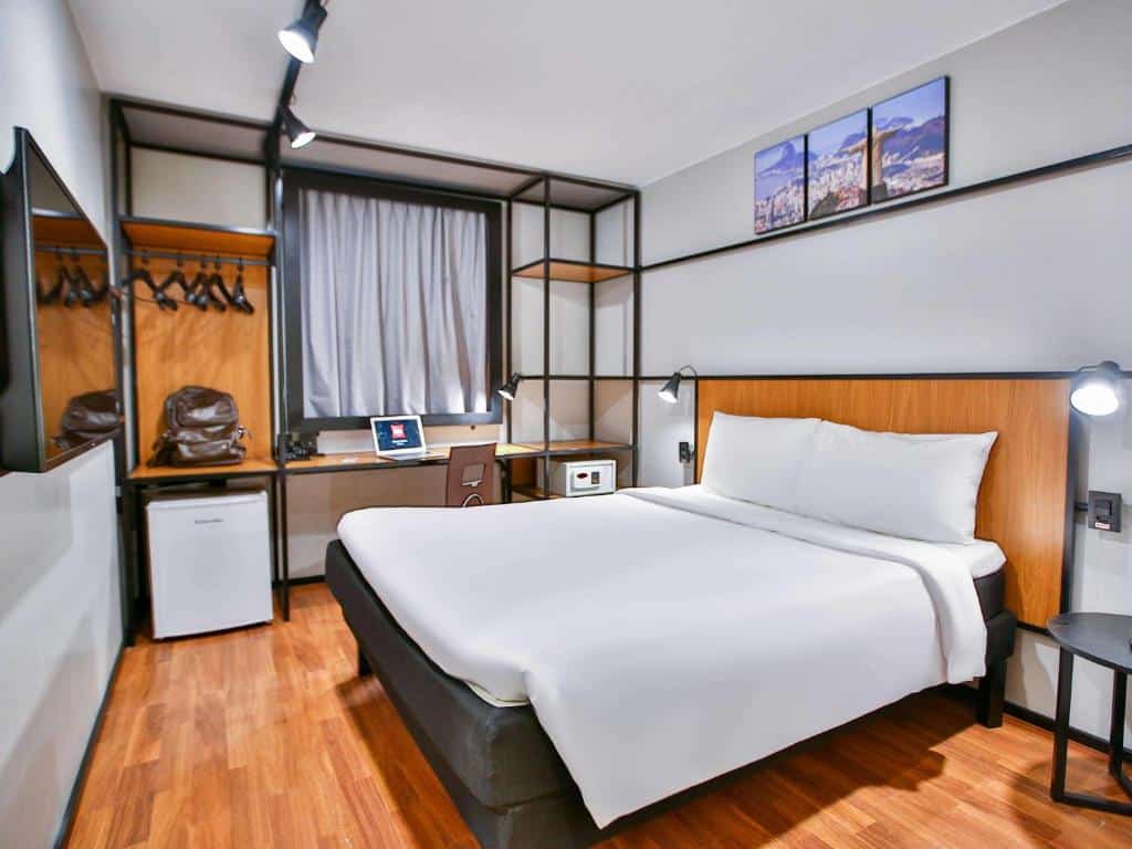Quarto do hotel com cama de casal branca, chão de madeira, escrivaninha, frigobar. cabides e uma tv, ilustrando post hotéis Ibis no Rio de Janeiro.