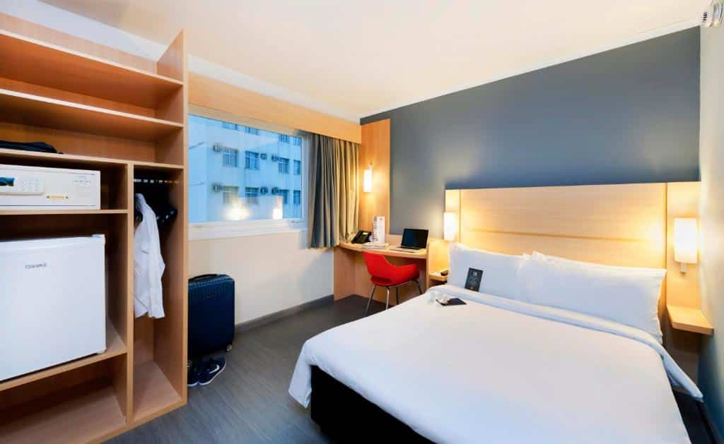 Quarto do hotel com uma cama de casal, guarda-roupa aberto com cabides, cofre e frigobar, escrivaninha com cadeira vermelha e uma janela de vidro, ilustrando post hotéis Ibis no Rio de Janeiro.