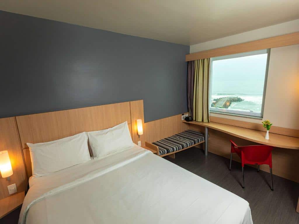 Quarto do hotel com uma cama de casal branca, parede cinza, escrivaninha com cadeira vermelha e uma janela de vidro com vista para a paisagem, ilustrando post hotéis Ibis no Rio de Janeiro.