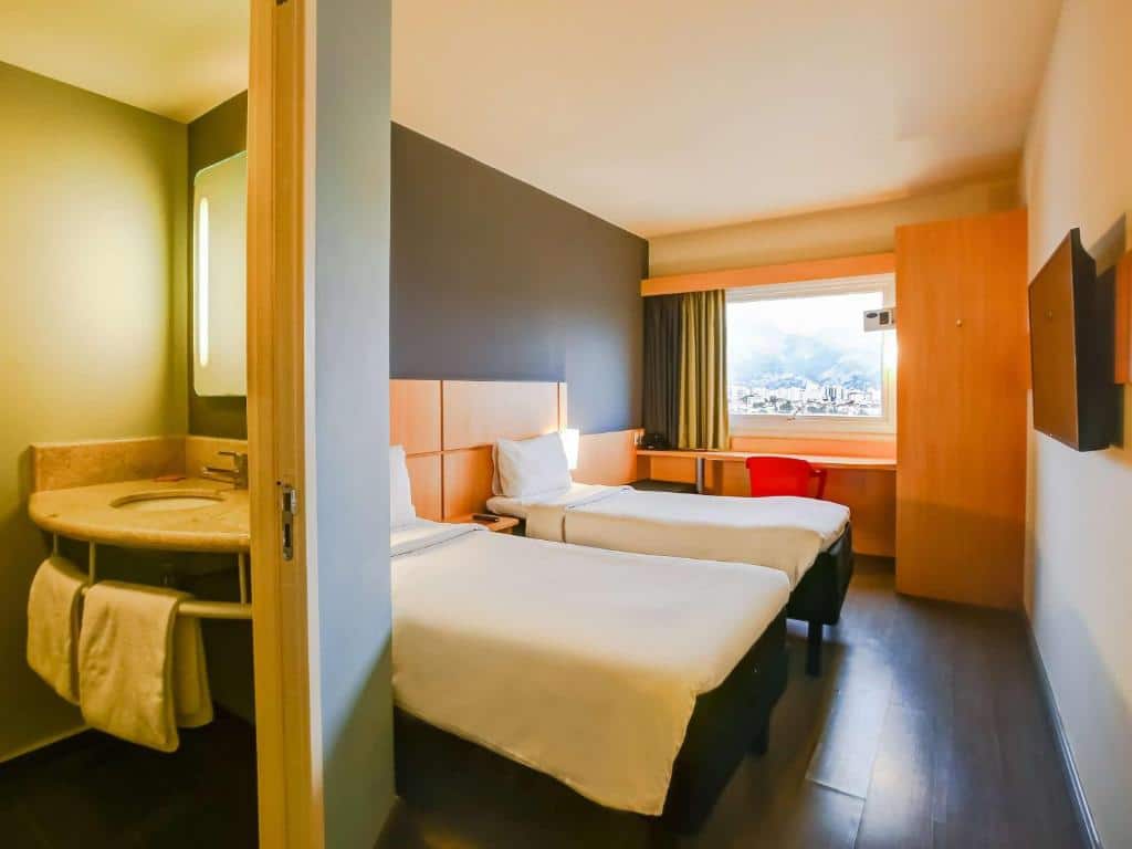 Quarto do hotel com duas camas de solteiro, uma tv, escrivaninha, janela de vidro e uma parte da imagem aparecendo a pia do banheiro, ilustrando post hotéis Ibis no Rio de Janeiro.