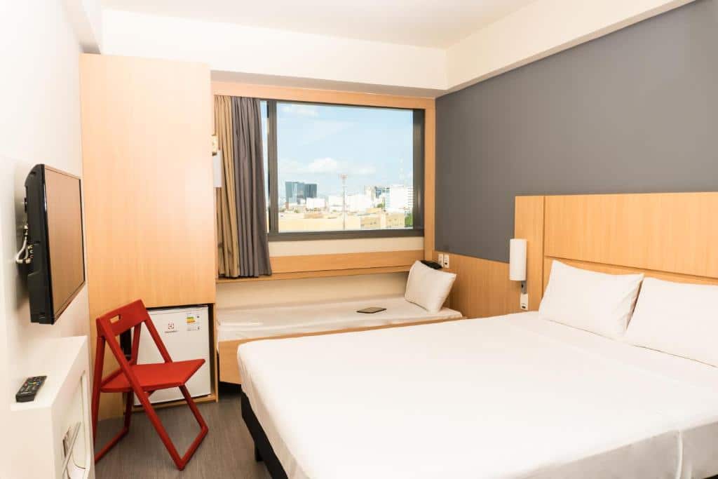 Quarto do hotel com uma cama de casal e uma de solteiro com lençóis brancos, janela de vidro, cadeira marrom, frigobar e uma tv, ilustrando post hotéis Ibis no Rio de Janeiro.