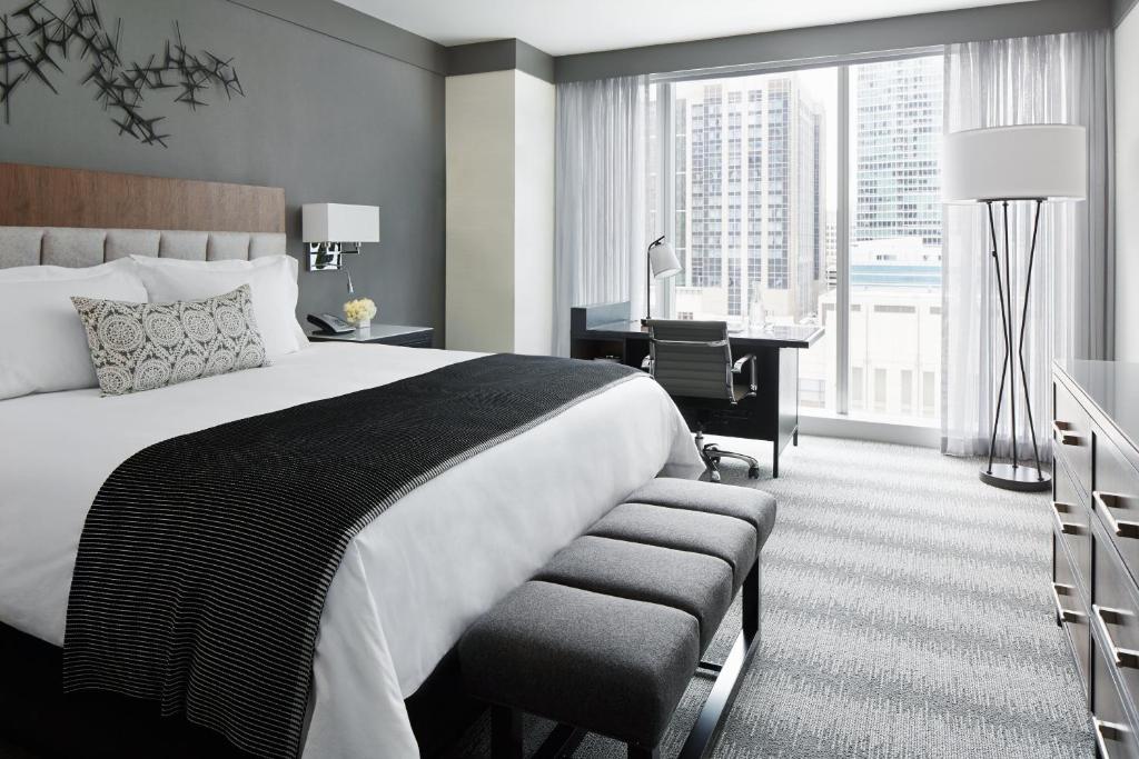 Quarto do Loews Chicago Hotel com cama de casal, cômoda do lado esquerdo com luminária, em frente a cama cômoda, do lado esquerdo uma mesa de trabalho e em frente a mesa janelas com cortinas brancas.