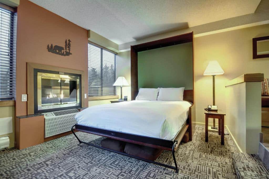 Quarto do Mountainside Lodge com cama de casal, uma lareira, duas janelas com persianas, chão de carpete e mesinhas com abajures em cima, para representar hotéis em Whistler