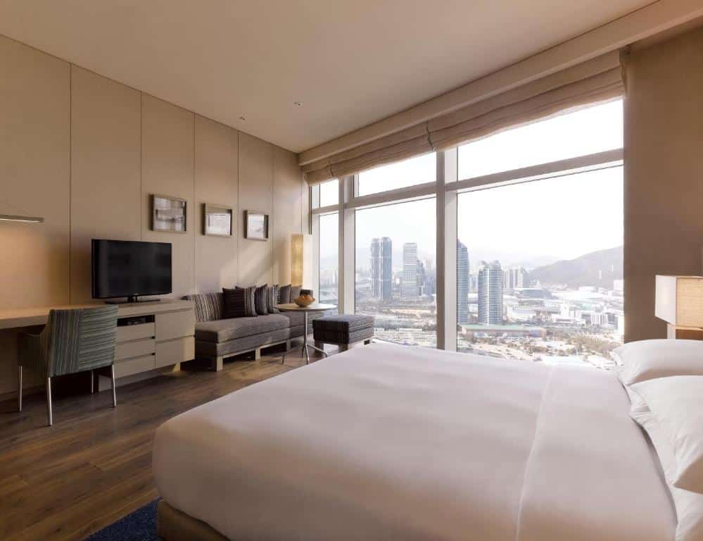 Quarto do Park Hyatt Busan com janela panorâmica com vista para a cidade, um sofá com almofadas, uma televisão e uma cama de casal