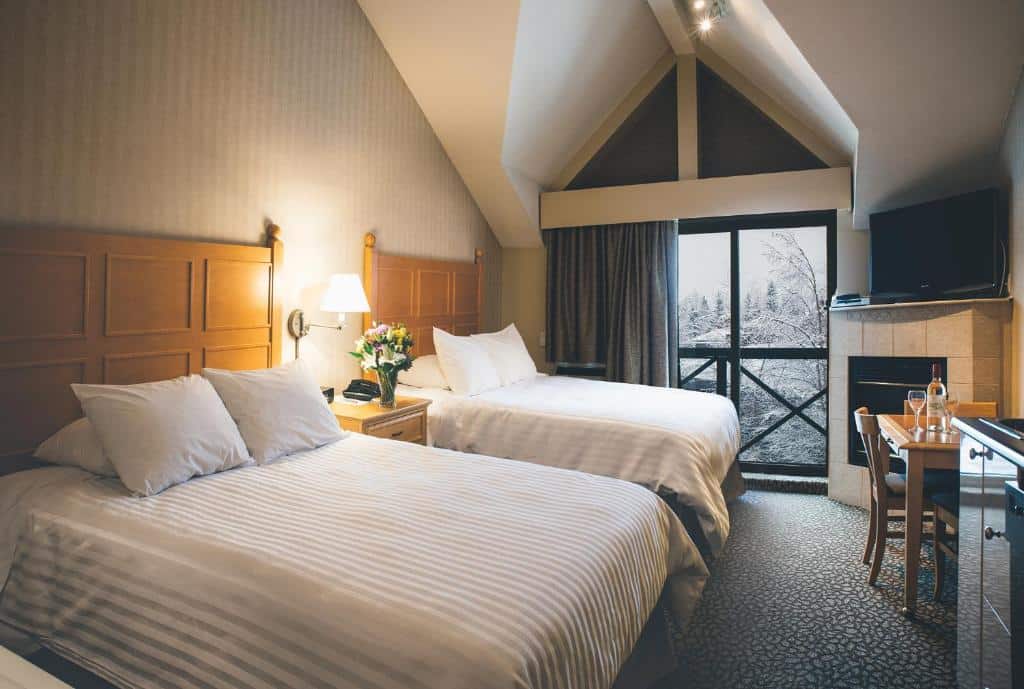 Quarto do Pinnacle Hotel Whistler com duas camas de casal, uma sacada com vista para as montanhas, uma lareira e uma televisão, além de uma mesa com quatro lugares, para representar hotéis em Whistler