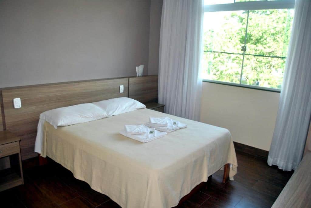 quarto da Pousada Alves em São Brás do Suaçuí com uma simples cama de casal ao centro e uma janela de vidro à direita, com vista para as árvores
