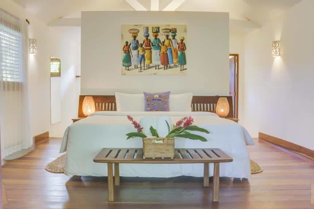 Quarto da Pousada Calypso com cama do casal ao centro do quarto com duas cômoda de madeiras ao lado da cama com luminárias e um banco de madeira no pé da cama. Representa resorts em Trancoso.