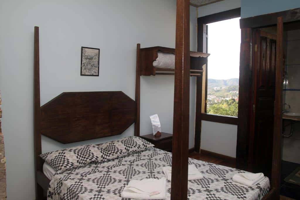 quarto da Pousada Gamarano com uma cama de casal em madeira, um armário ao lado, uma janela ampla aberta com vista para o vale mineiro.