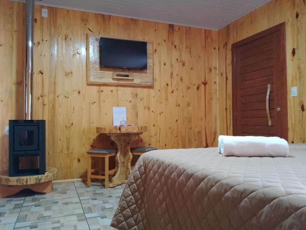interior do chalé em Cambará do Sul com uma lareira à esquerda, ao lado de uma mesa pequena e redonda de madeira com dois banquinhos, embaixo de uma televisão de tela plana e em frente a cama de casal.