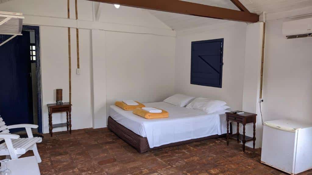 Quarto da Pousada Roda D’água com frigobar do lado direito, do lado esquerdo cama de casal com uma cômoda de madeira ao lado.