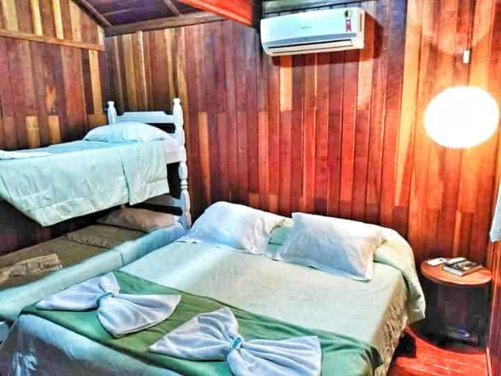 Quarto do Pousada Sabiá com cama de casal do lado direito com uma cômoda de madeira redonda e do lado esquerdo uma cama beliche.