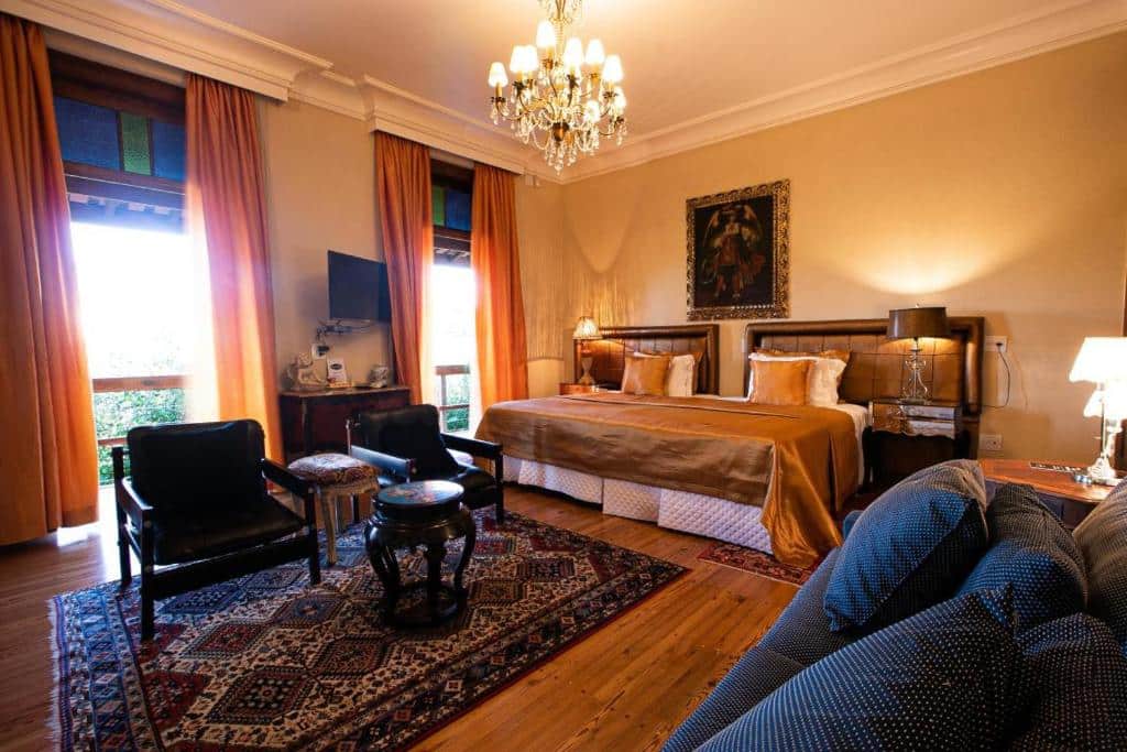 Quarto do Villa Maria Pousada de Charme com cama ampla, duas cômodas ao lado da cama com luminária, ao centro do quarto duas poltronas pretas com mesinha preta no centro e sofá de dois lugares azul em frente as poltronas.