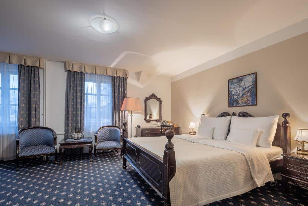 Quarto espaçoso do Pytloun Old Armoury Hotel Prague com carpete azul marinho com detalhes em branco, duas janelas com cortinas, uma cama de casal, duas mesinhas de cabeceira com abajures, uma penteadeira com um espelho e duas poltronas