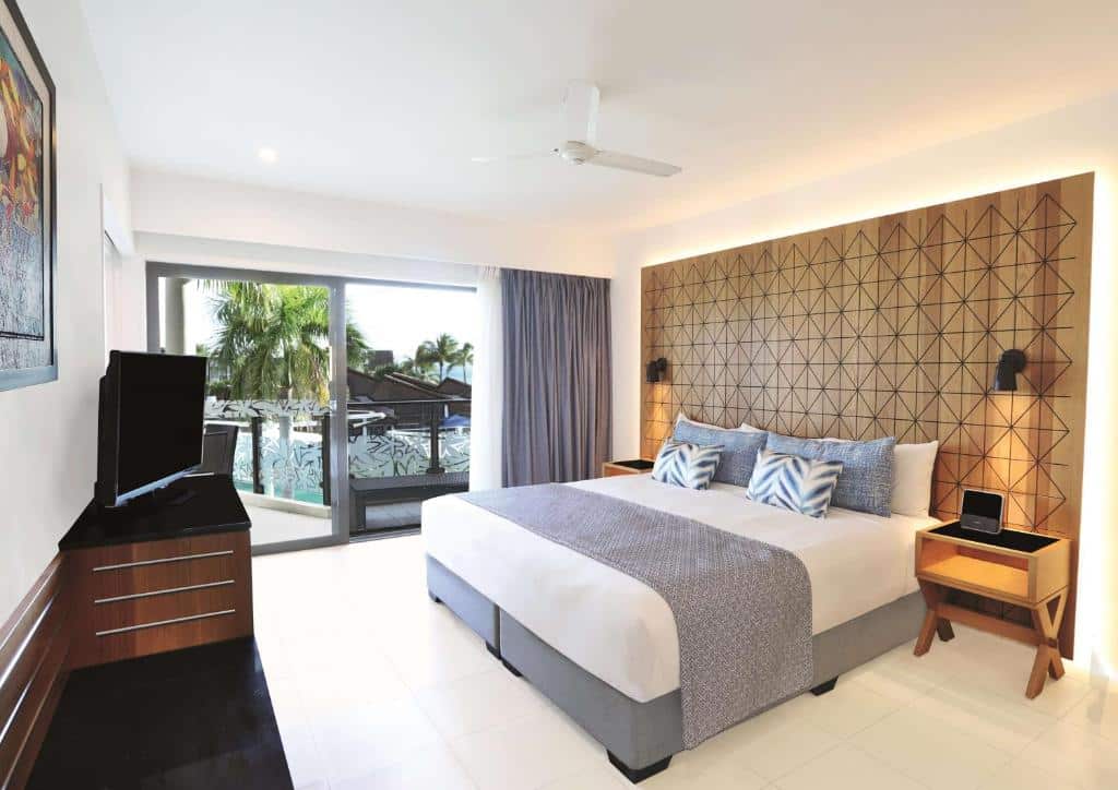 Quarto do Radisson Blu Resort com cama de casal, com duas cômodas de madeira ao lado e em frente a cama cômoda de madeira com TV em cima e do lado esquerdo portas de vidro com saída para o térreo.