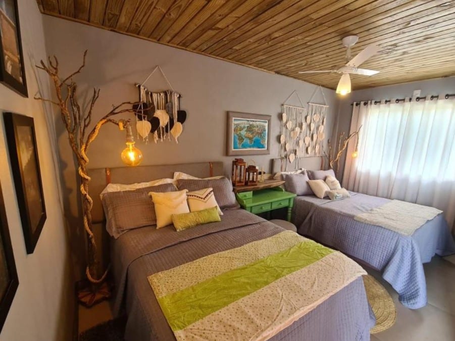 quarto do hotel refúgio dos coiotes em Cambará do Sul; O espaço mostra vários enfeites artesanais pendurados nas paredes do fundo, há duas camas de casal dividindo o espaço com uma pequena mesa verde entre as camas.