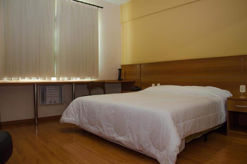 quarto do Serra Palace Hotel em Ouro Branco com uma cama de casal e uma mesa de madeira ampla, situado logo abaixo da janela que está fechada por uma cortina branca, com blackout