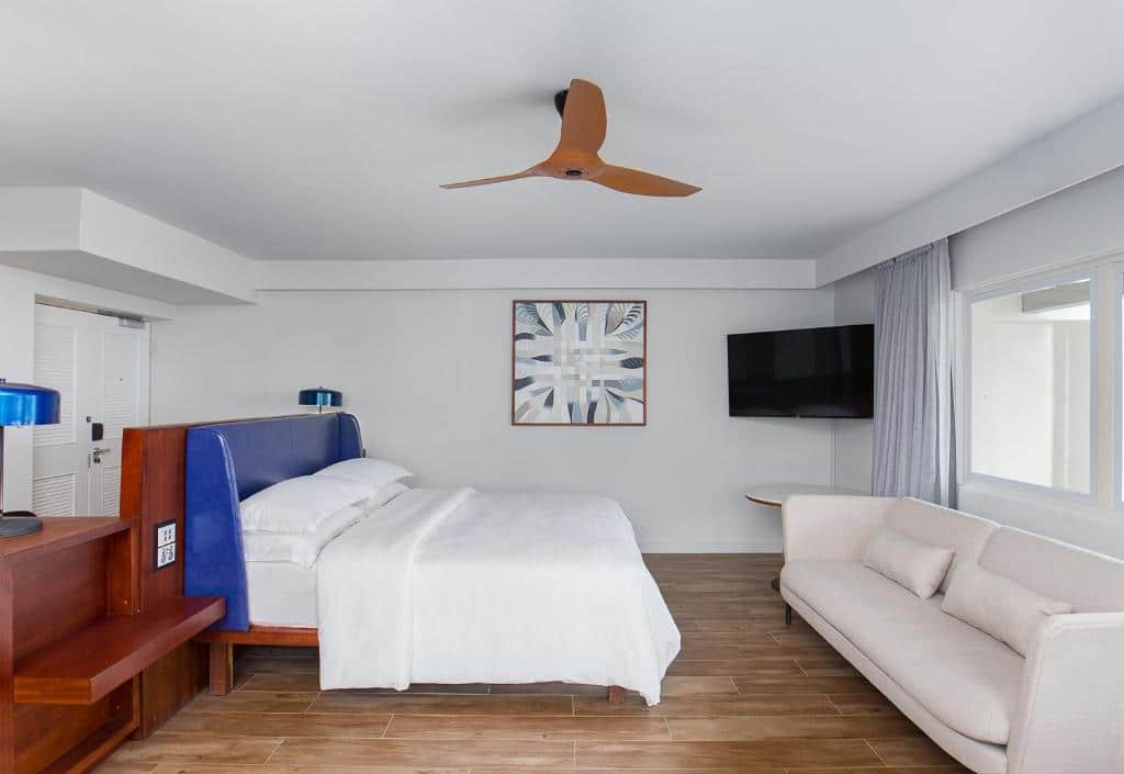 Quarto do Sheraton Fiji Golf & Beach Resort com cama de casal, ventilador no teto em cima da cama, sofá de dois lugares em frente a cama com TV presa na parede com mesa redonda abaixo.