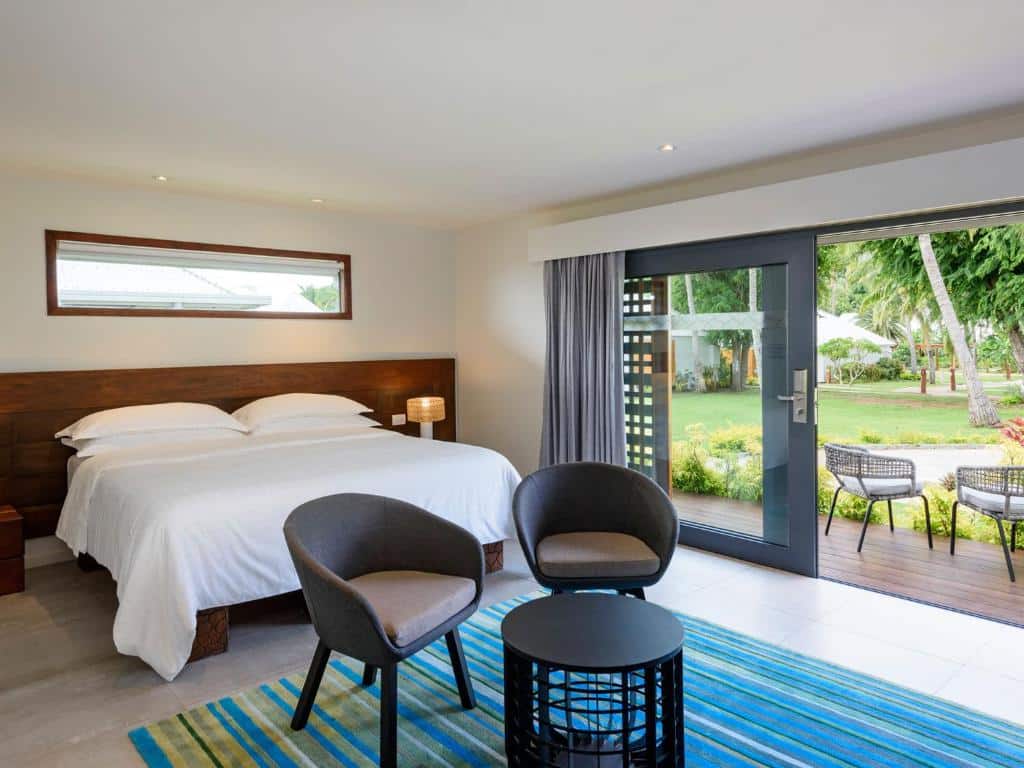 Quarto do Sheraton Resort & Spa, Tokoriki Island com cama de casal, duas cadeiras pretas no pé da cama com mesinha redonda preta, do lado esquerdo portas de vidro com acesso ao térreo.