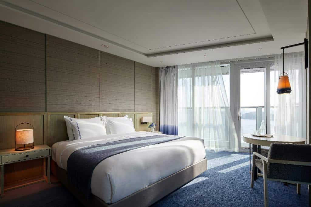 Quarto do Signiel Busan com uma sacada com vista para o mar, carpete azul marinho no quarto, uma cama de casal, uma cabeceira com luminárias e uma mesa redonda com uma cadeira