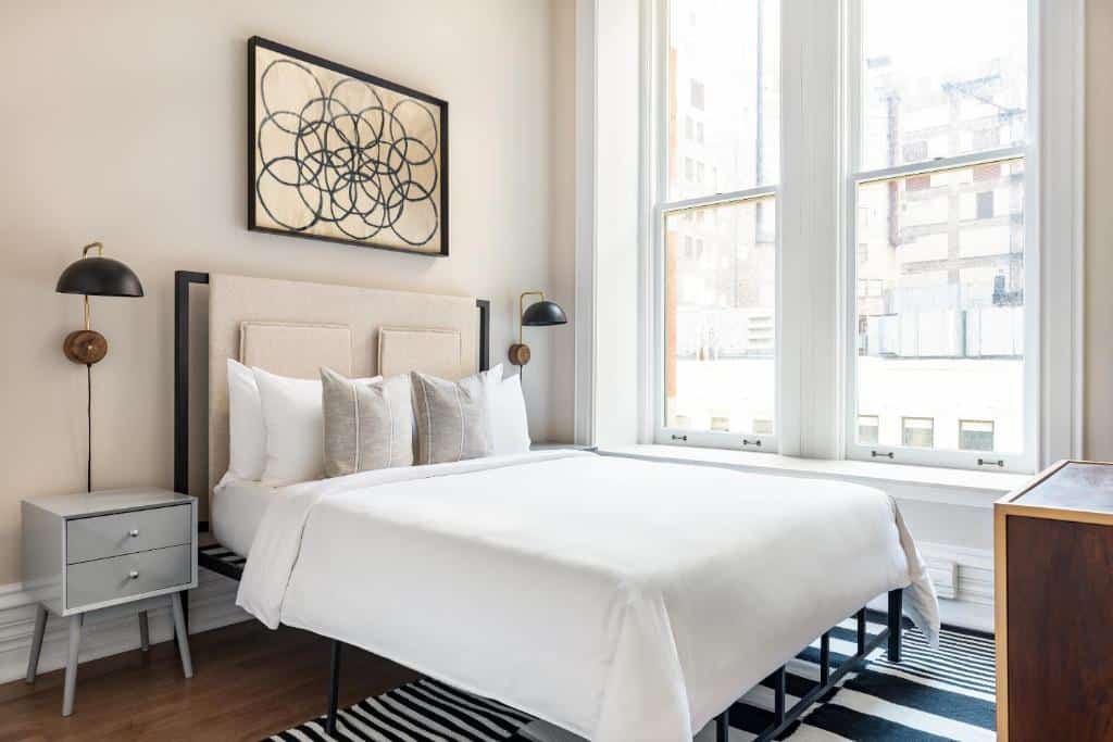 Quarto do Sonder Jewelers Row com cama de casal, duas cômodas cinza claro com luminária, janelas do lado esquerdo. Representa hotéis em Chicago.