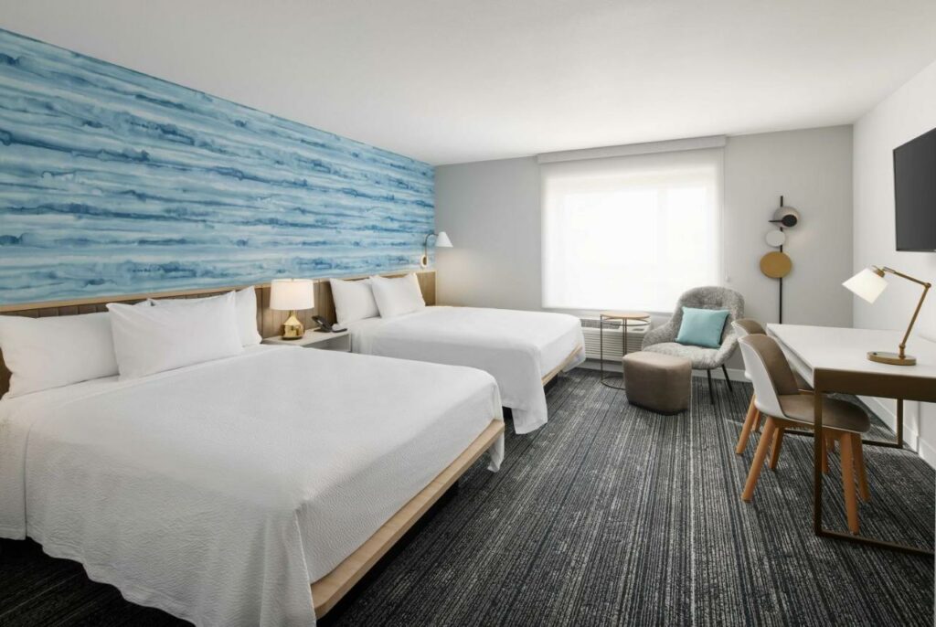 Quarto do TownePlace Suites by Marriott Tampa Casino Area com uma cama de casal, uma de solteiro, um carpete cinza com branco, uma janela com cortinas, uma bancada de trabalho com duas cadeiras e uma poltrona
