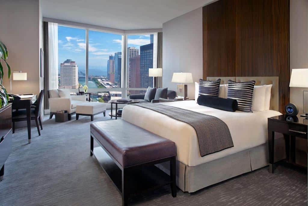 Quarto do Trump International Hotel & Tower com cama de casal, duas cômoda ao lado com luminária, do lado esquerdo sofá cinza com mesa de vidro e outro sofá branco perto das janelas panorâmicas de vidro.