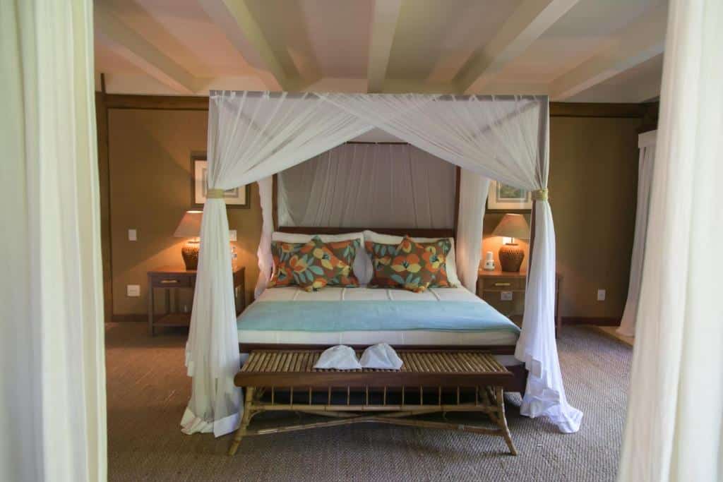 Quarto do Pousada Tutabel com cama de casal ao centro da cama, com duas cômodas de madeira com luminárias de cada lado da cama e um banco de madeira ao pé da cama.