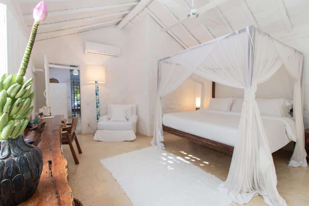 Quarto do Uxua Casa Hotel & Spa com cama de casal do lado direito, poltrona branca do lado esquerdo da cama, com mesa de madeira rústica em frente a cama.
