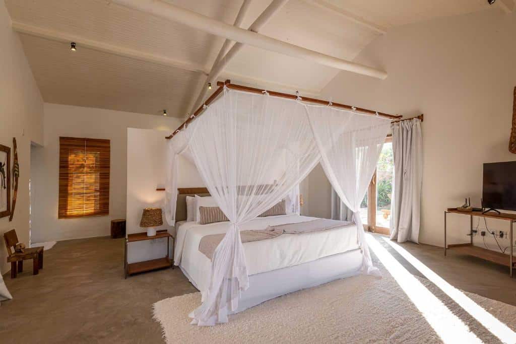 Quarto do Villa dos Nativos Boutique Hotel com cama de casal ao centro do quarto, uma cômoda de madeira com luminária, portas de vidro com cortinas brancas do lado esquerdo do quarto, com uma cômoda de madeira com TV em cima.
