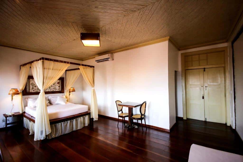 Quarto da Pousada Portas da Amazônia com cama de casal do lado esquerdo com mosqueteiro, duas cômodas de madeira com luminária e uma mesa com duas cadeiras no centro do quarto.