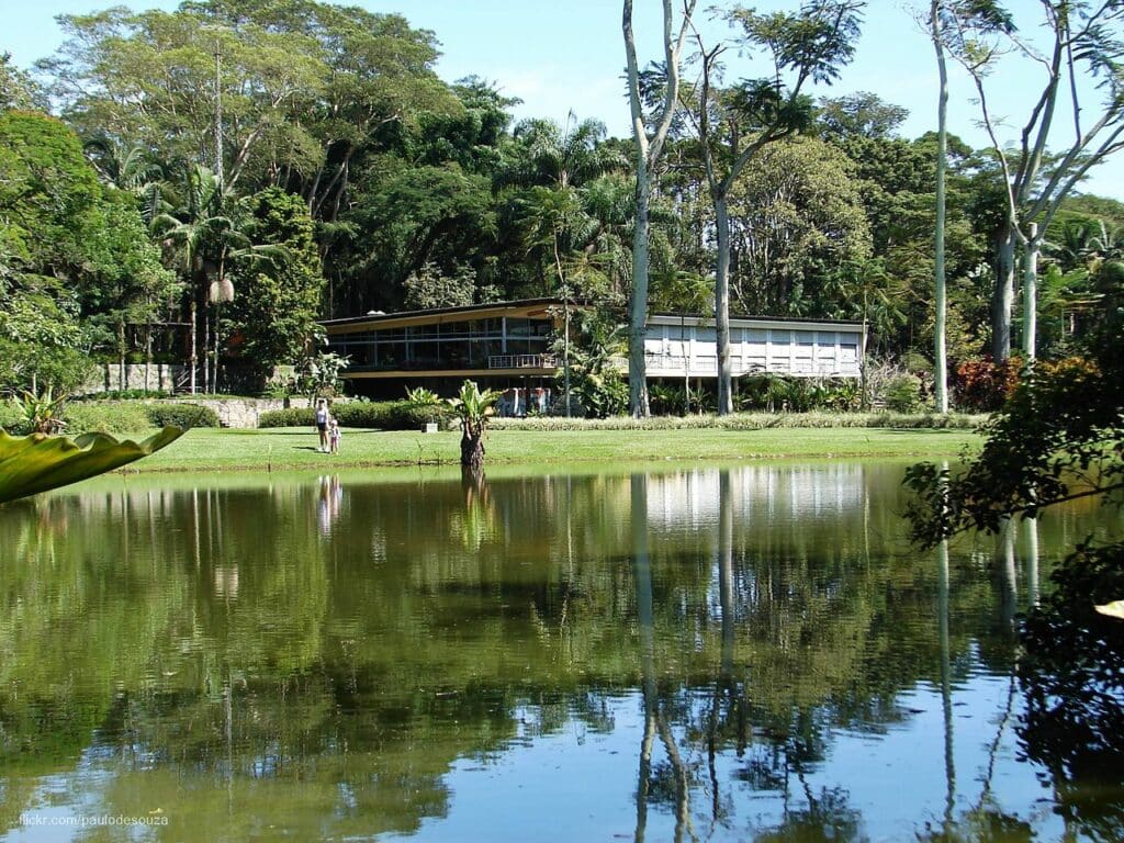 Vista da Residência Olivo Gomes, atrás do lago do Parque da Cidade, envolta por árvores, com uma pessoa e uma criança ao longe, olhando em direção à câmera