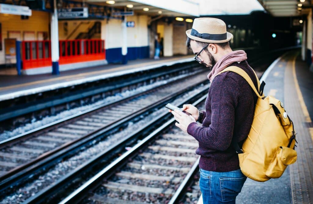 Homem usando um chapéu e mochila amarela nas costas mexendo no celular com as duas mãos. Ele está na beira dos trilhos de trem em uma estação no Reino Unido