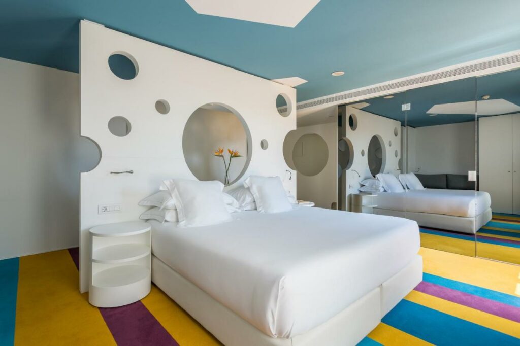 Quarto do Room Mate Pau, uma das recomendações de hotéis boutique em Barcelona. A cama tem lençol e travesseiros brancos e está encostada em uma divisória de ambientes branca que lembra um queijo esburacado. Há mesinhas de cabeceira brancas dos dois lados da cama, e o chão do quarto é listrado das cores amarela, azul e rosa. Há um espelho que ocupa a parede lateral.