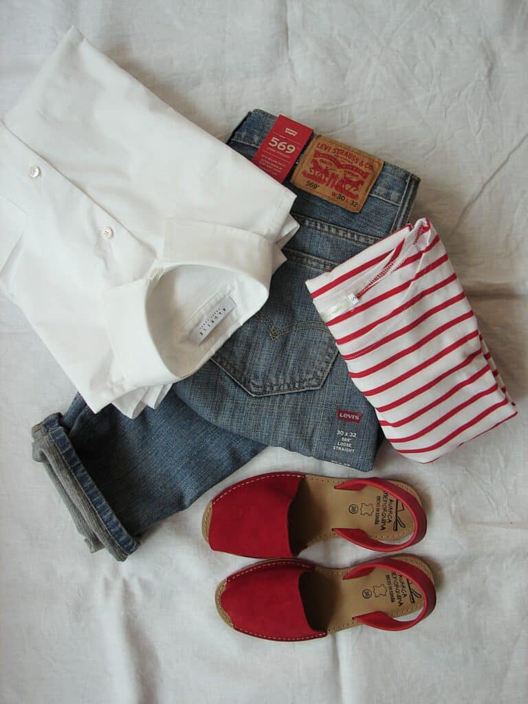 Camisa branca, blusinha branca com listras vermelhas, calça jeans levis da cor azul médio dobrados, ao lado de um sapato sem salto, fechado na frente, e vermelho