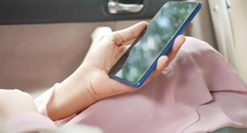 mão feminina branca segurando um celular smartphone dentro de um carro. É possível enxergar um casaco rosa deixado sobre suas pernas, onde a mão está apoiada com o celular