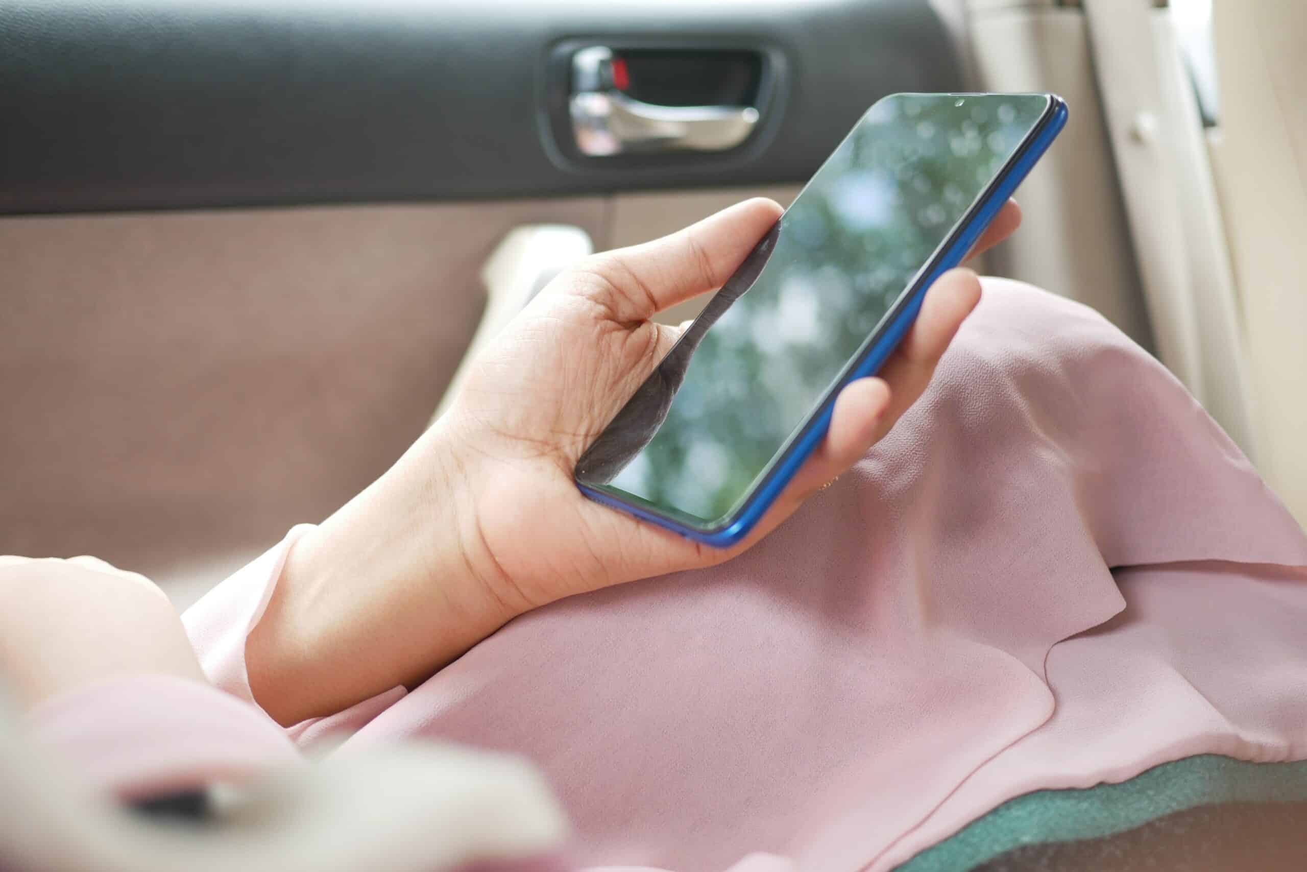 mão feminina branca segurando um celular smartphone dentro de um carro. É possível enxergar um casaco rosa deixado sobre suas pernas, onde a mão está apoiada com o celular