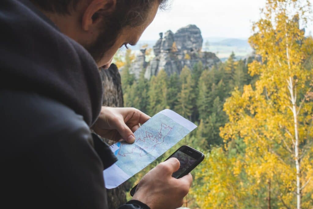 Homem usando blusa de frio cinza escuro segurando um mapa com uma mão e um celular com a outra. Ao fundo tem uma paisagem com árvores altas e uma rocha grande no  horizonte