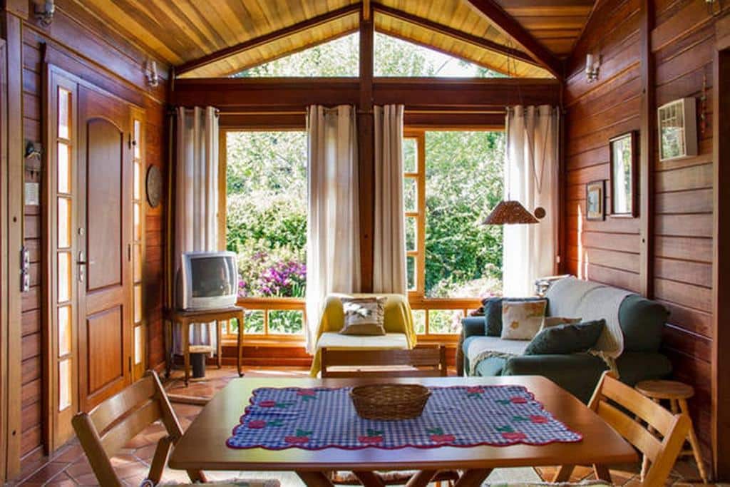 Sala de estar com amplas janelas em um dos chalés da Vila Báltica Chalés, inteiro de madeira, com sofás, uma vista para o verde, e uma mesa de madeira com duas cadeiras